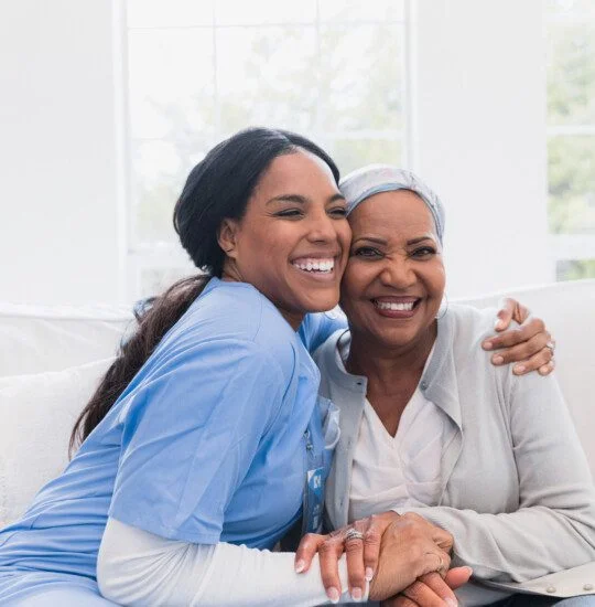 senior woman and her caregiver smile and hug while seated on a couch