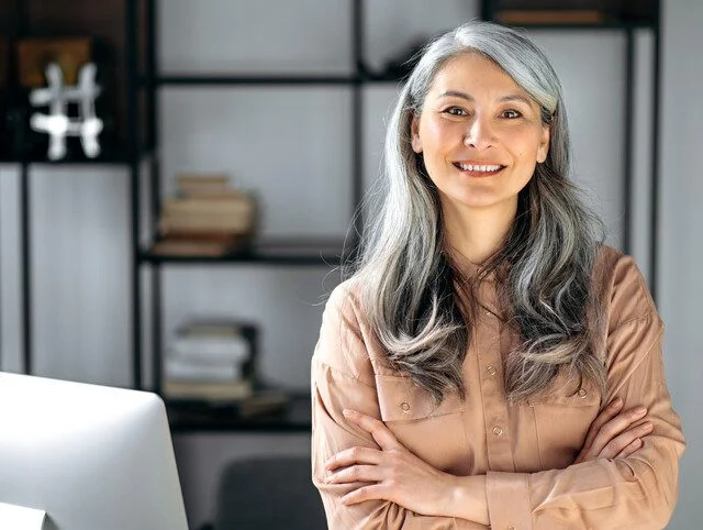 Portrait of a successful confident mature gray-haired lady, business woman, ceo or business tutor, standing in the office with arms crossed, looking and friendly smiling into the camera