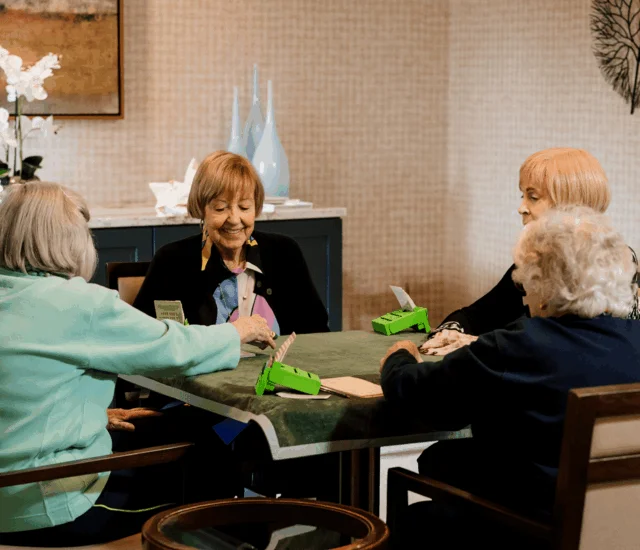 Four women sit around table playing cards with green card holder and tissue box.