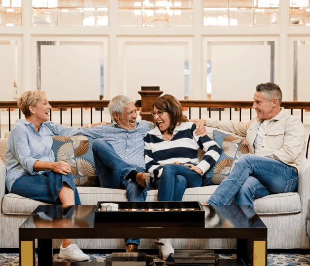 Four people laughing on a couch with patterned pillows and a coffee table below them.
