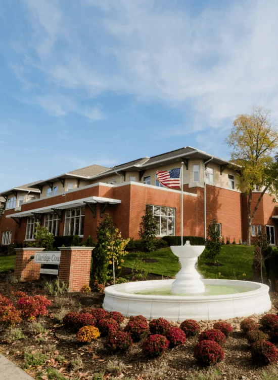 A brick building with an American flag and a white fountain surrounded by autumn foliage under a blue sky.
