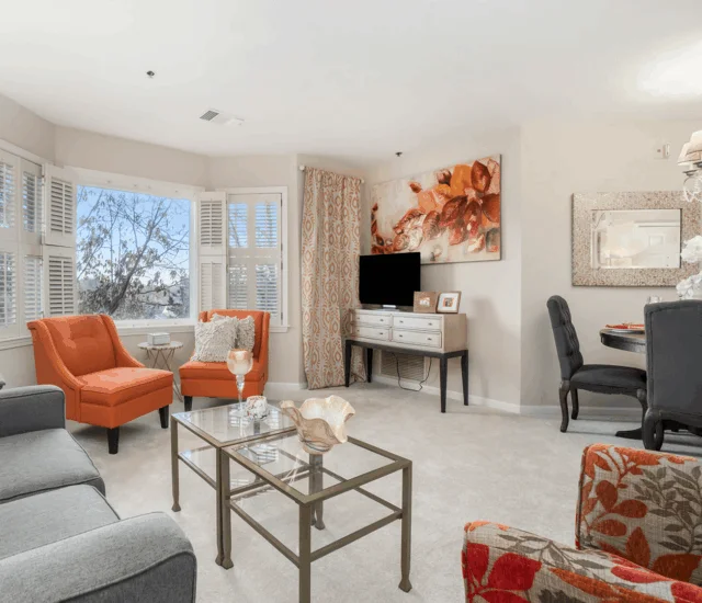 A cozy living room with orange chairs, gray sofa, glass coffee table, and abstract art on wall.