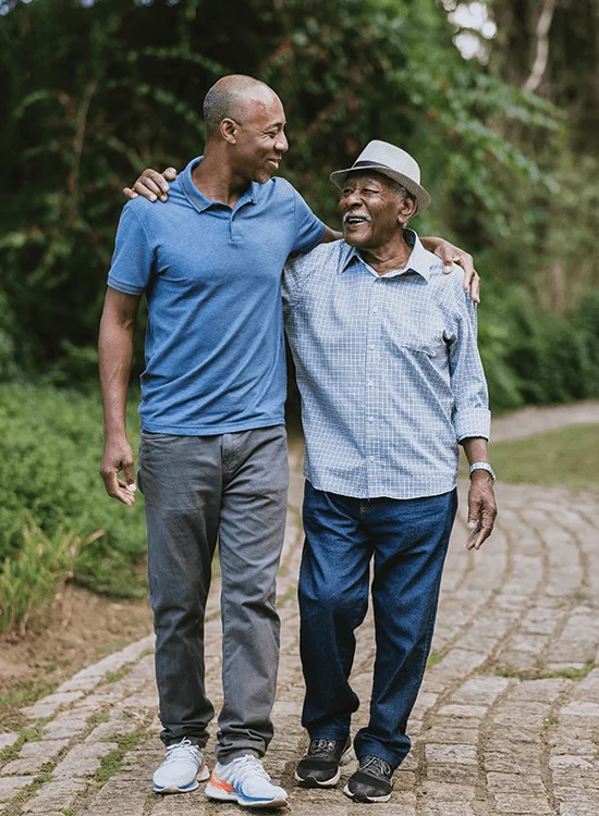 Two men walk together on a stone path surrounded by greenery.