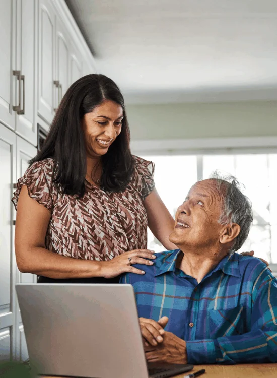 A woman smiles as she helps an older man with his laptop at home.