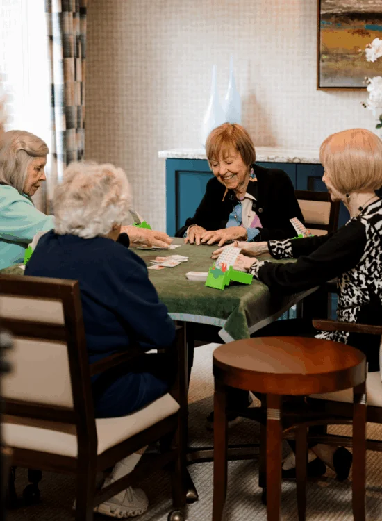 Four women play cards at a table with green cloth and wooden chairs indoors.