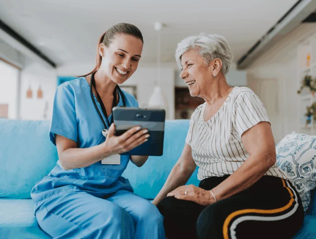 Nurse and senior share smile with tablet on blue couch.