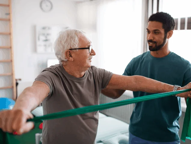 A therapist assists an elderly man with resistance band exercise indoors.