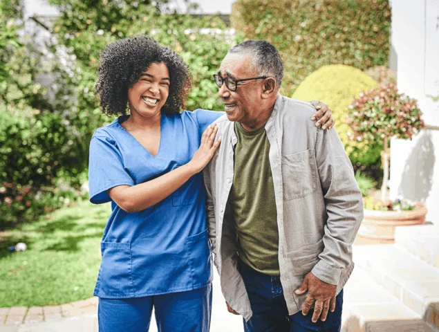 A caregiver and senior share a joyful moment outdoors near greenery.