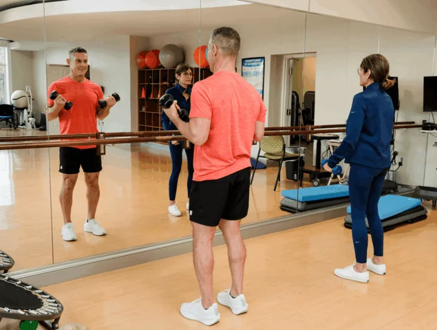 Man and woman exercising with dumbbells near mirrors in gym setting.