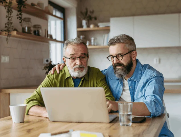 Two men with beards and glasses use a laptop together at home.