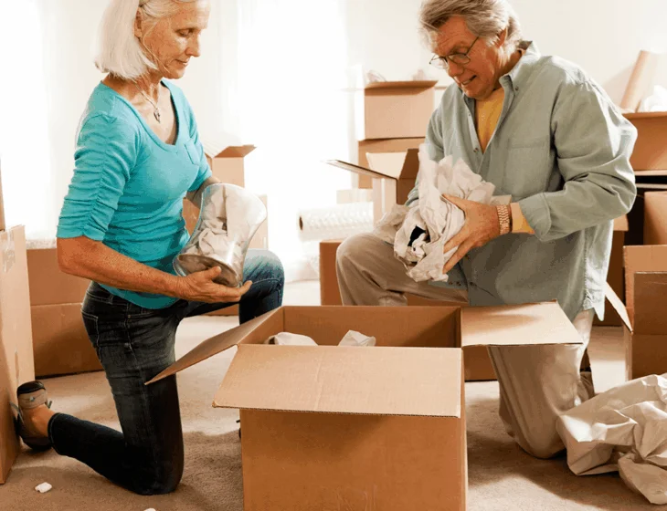 Senior couple unpacking boxes on floor with items and tissues around them.