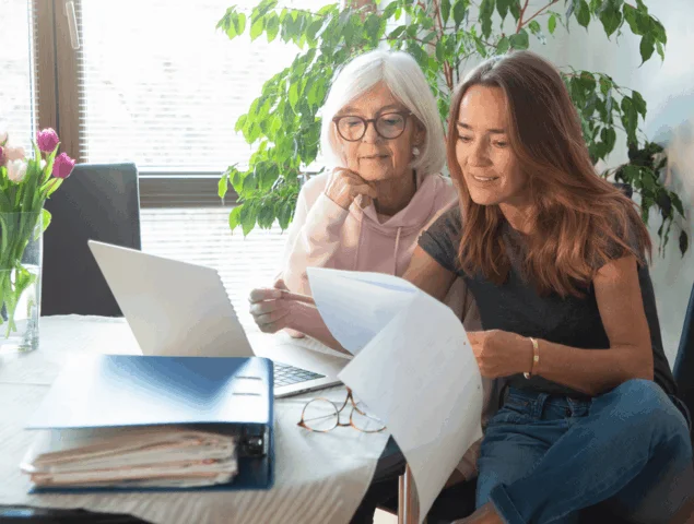 Two women review documents and use laptop at table with flowers nearby.