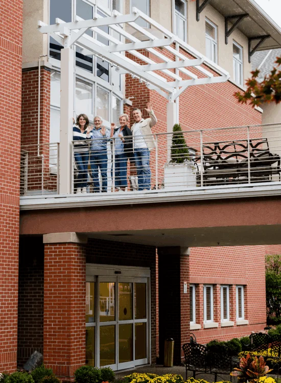 Four people wave from a balcony with brick and glass architecture below.