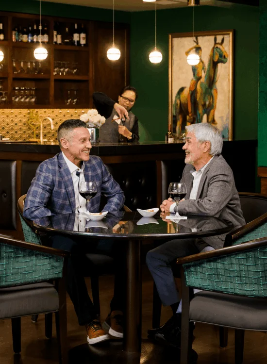 Two men sit at a table with wine glasses and bowls, smiling as they converse in a cozy bar setting.