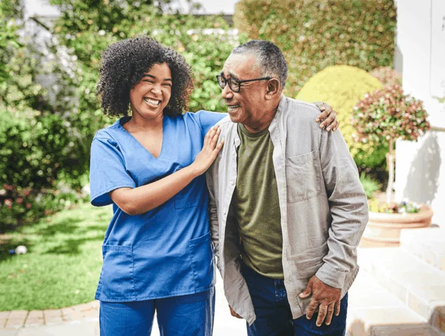 A caregiver and senior share a joyful moment outdoors near greenery.