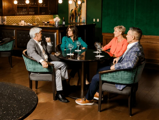 Four people sit around a table with wine glasses, engaged in conversation in an elegant room.