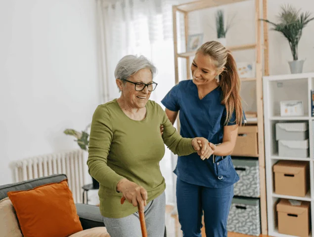 A caregiver assists an elderly woman with walking using a cane indoors.