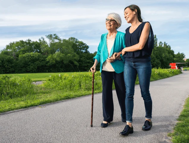 Two women walk together on a paved path surrounded by greenery under a partly cloudy sky.