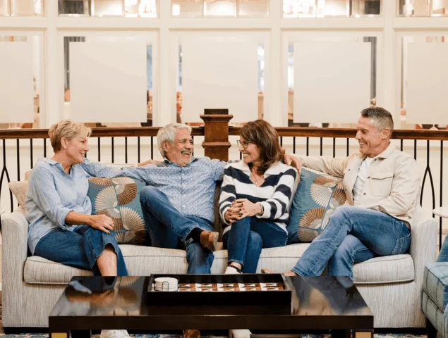 Four people sit on a couch laughing and chatting together indoors.