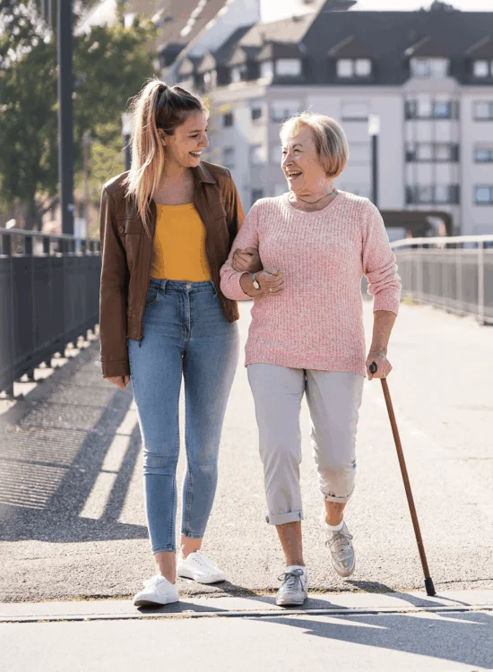 Two individuals walk together on a sunny day; one uses a cane while smiling and talking.