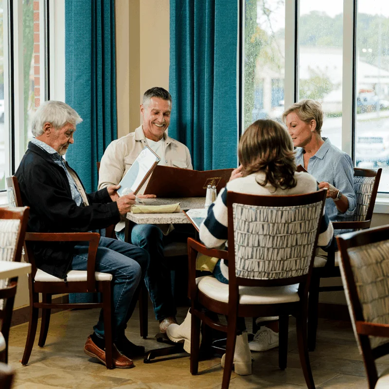 Four people sit at a table with menus and drinks, enjoying conversation near large windows.