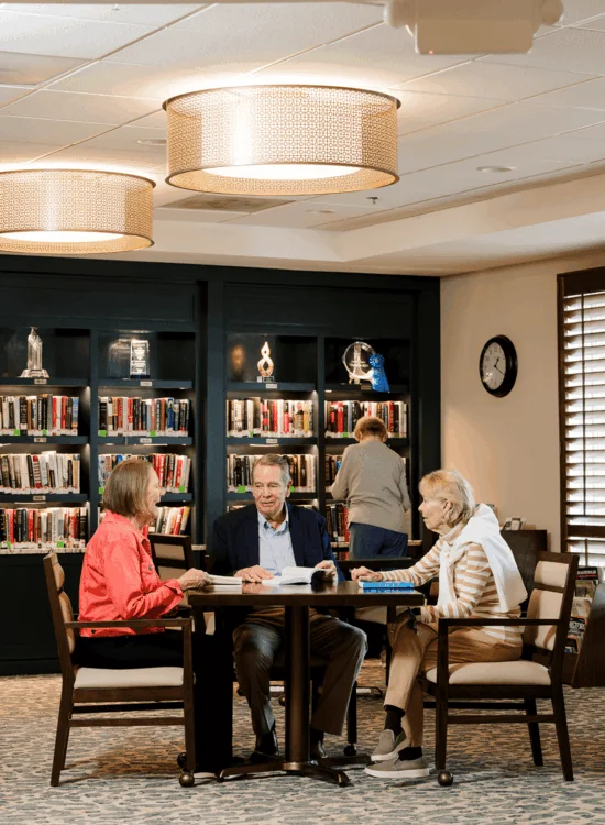 Three people sit at table with books, discussing in library setting.