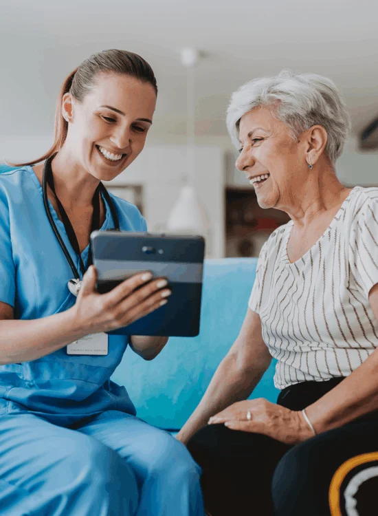 A nurse and elderly woman share a smile while looking at a tablet together indoors.