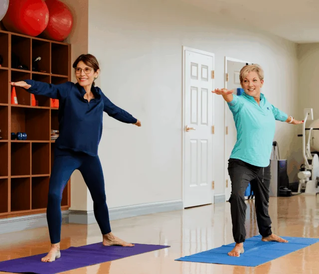 Two women practice yoga on mats indoors with exercise equipment and storage shelves nearby.