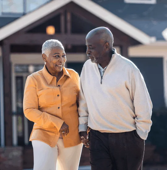 A smiling couple walks hand-in-hand outside their home on a sunny day.