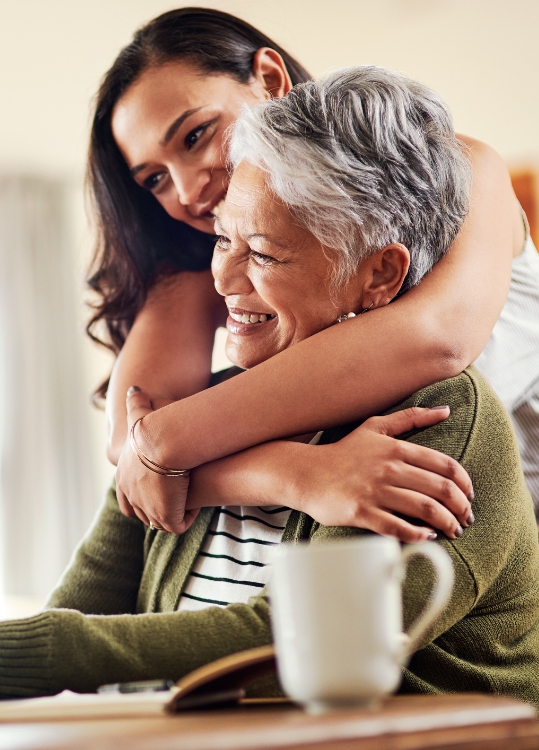 adult daughter leans over the back of her grandmother's chair to hug her from behind, both smiling