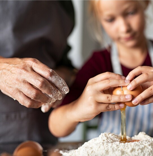 grandparent shows granddaughter how to crack egg while making bread