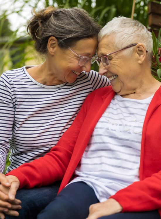 senior woman and her adult daughter snuggle close outdoors, touching foreheads