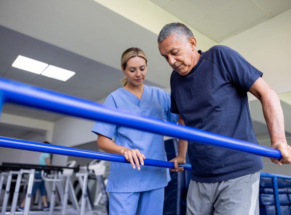 senior man uses bars to hold himself up with the aid of a physical therapist during a rehabilitation session