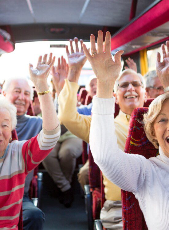 group of seniors smile and raise their hands up seated in a bus