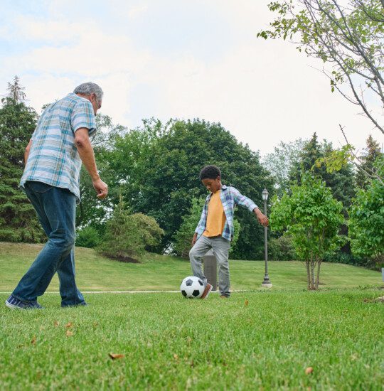 grandfather and grandson play soccer in the grass outside of his senior living community