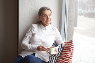 senior woman on a window seat inside her home holds a cup of tea and looks sadly out of the window