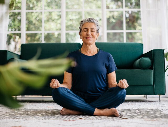 senior woman sits on the floor of her home in a meditative seated yoga position, smiling with eyes shut