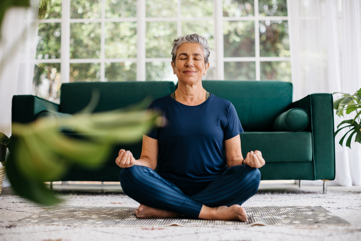 senior woman sits on the floor of her home in a meditative seated yoga position, smiling with eyes shut
