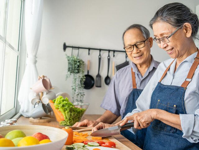 senior couple wearing matching aprons smile while prepping vegetables for a meal in their kitchen