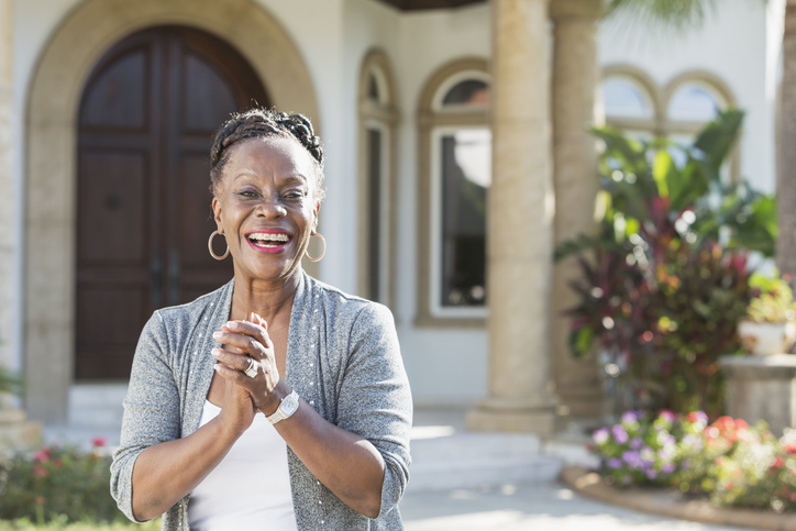 senior African American woman smiles and clasps her hands together while standing in front of her stunning and lavish home entrance