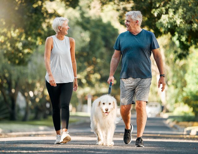 senior couple in athletic clothing smiles and walks their dog on a scenic path