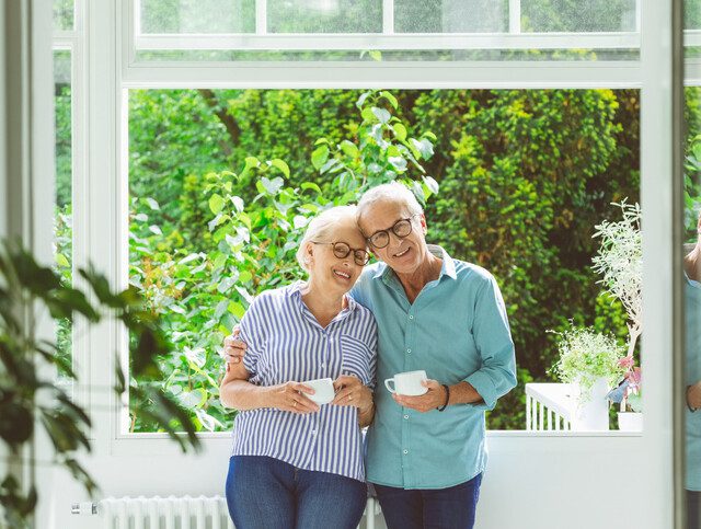 senior couple holding coffee cups smile and lean their heads together, standing in front of an open window with beautiful landscaping visible