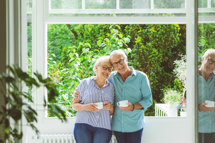senior couple holding coffee cups smile and lean their heads together, standing in front of an open window with beautiful landscaping visible