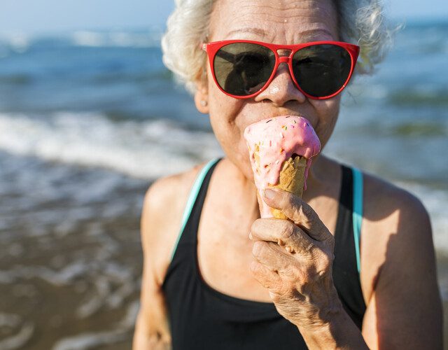 senior woman in bathing suit and red sunglasses eats ice cream cone at the beach