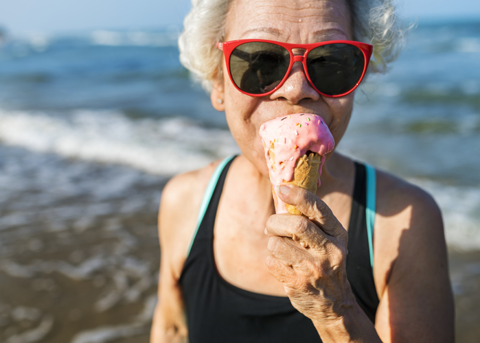 senior woman in bathing suit and red sunglasses eats ice cream cone at the beach