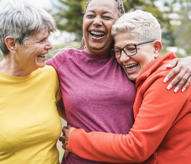 three close senior female friends in athletic clothing laugh and hug outdoors