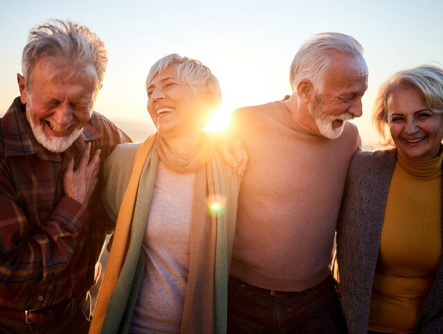 group of three well-dressed seniors laugh and wrap their arms around each other while walking, backdropped by the sun