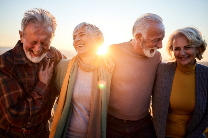 group of three well-dressed seniors laugh and wrap their arms around each other while walking, backdropped by the sun
