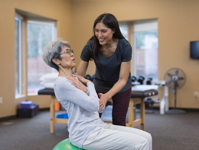 Senior woman practices balance exercise on an exercise ball with the help of a physical therapist