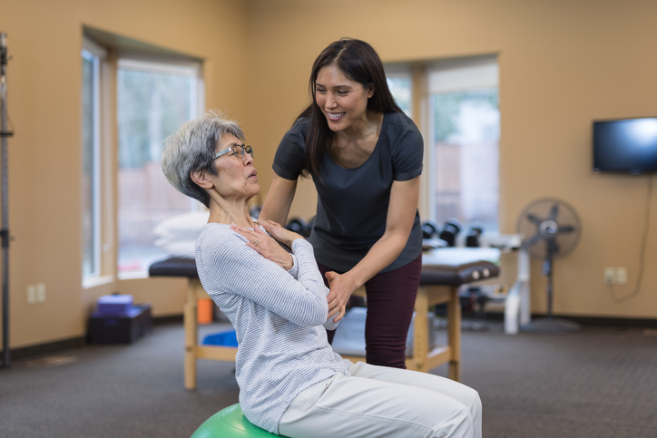 Senior woman practices balance exercise on an exercise ball with the help of a physical therapist